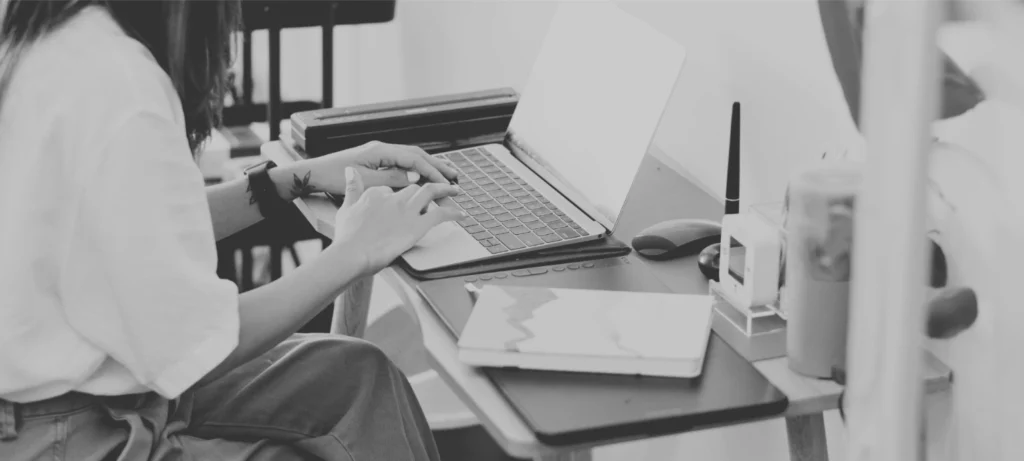 nainen istuu työpöydän ääressä ja kirjoittaa läppärillä. Pöydällä on toimistotarvikkeita