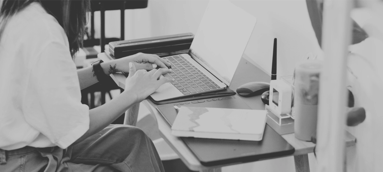 nainen istuu työpöydän ääressä ja kirjoittaa läppärillä. Pöydällä on toimistotarvikkeita